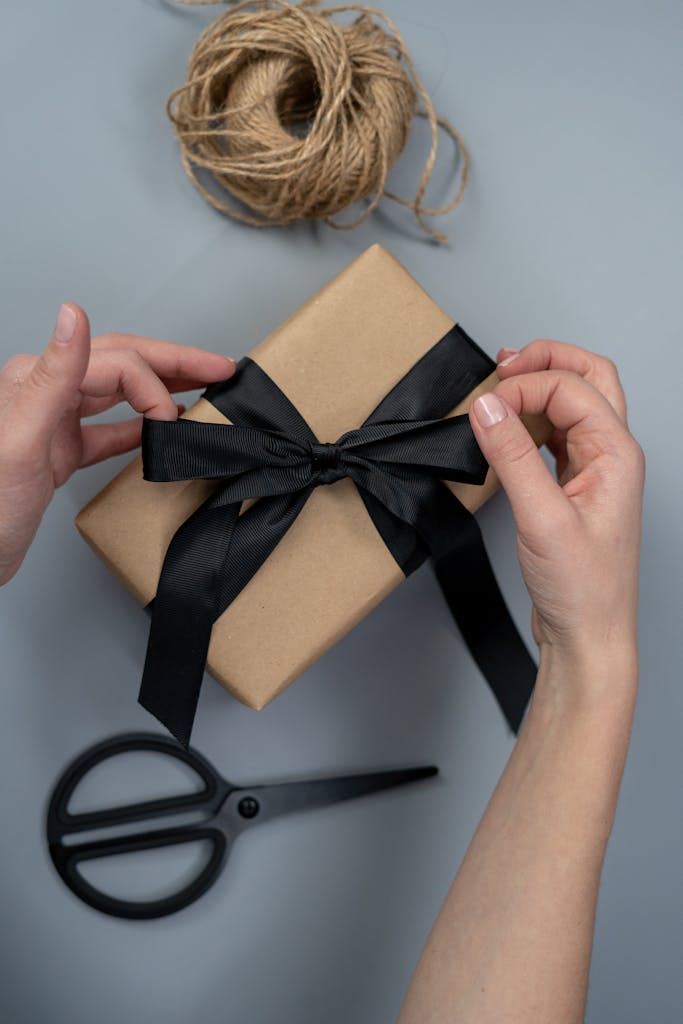 Hands tying a black ribbon on a craft paper gift box, next to scissors and twine.